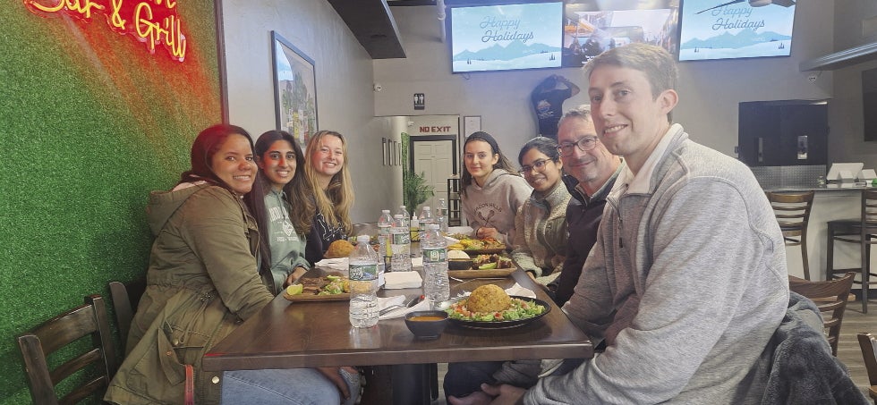 A group of seven people sit around a rectangle high top chair in a restaurant with plates of food in front of them.